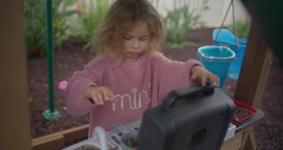 Young girl plays pretend kitchen outdoors, stirring leaves and bubbles in a backyard playhouse
