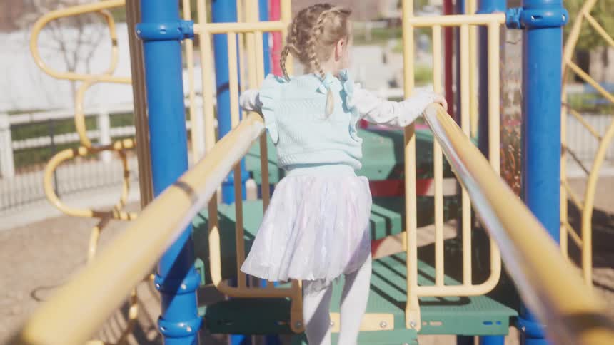 A little girl with pigtail braids carefully walks across a colorful playground structure in slow motion on a sunny day