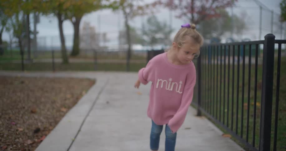 A playful little girl in an oversized pink sweater dances in slow motion on a walkway at a park
