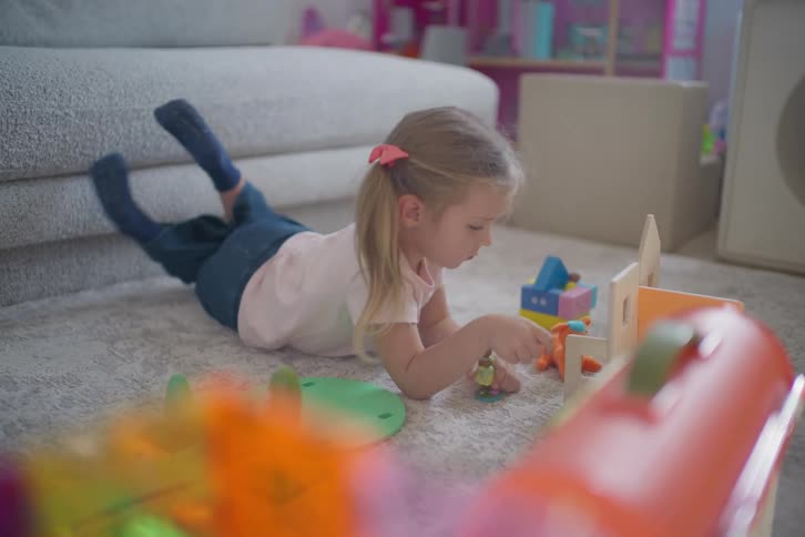 Young girl lies on the living room carpet playing with toy figures and a dollhouse in soft natural light