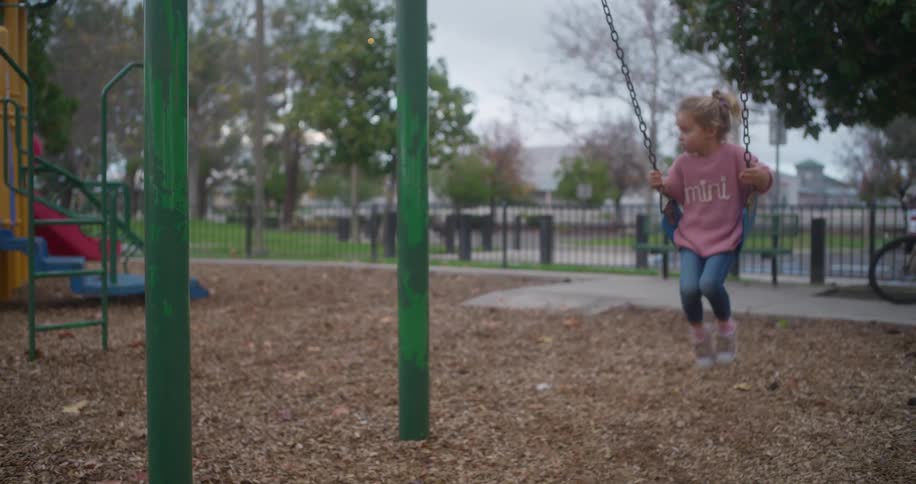 A cute little girl in a pink sweater swings gently back and forth in slow motion at an outdoor park playground