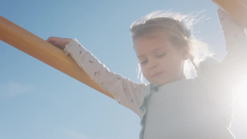A little girl with windblown hair plays on a yellow jungle gym as the bright sun flares behind her against a clear blue sky