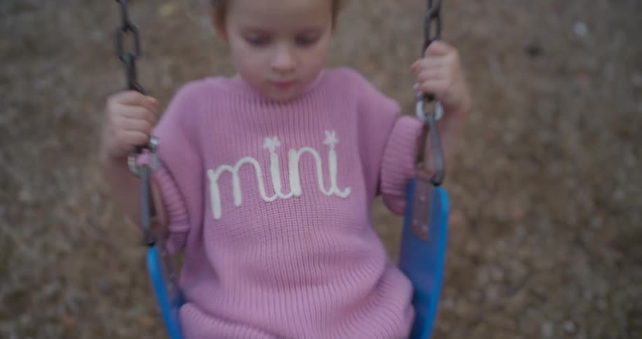 A cute little girl in a pink sweater swings back and forth in slow motion as the camera focuses on her pensive face
