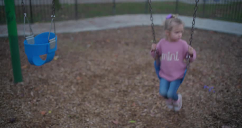 Slow motion shot of a cute toddler girl in a pink sweater swinging thoughtfully on a swing set at the playground