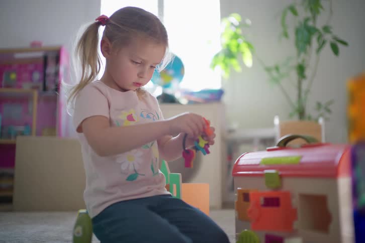 A young blonde girl with pigtails kneels on the floor, intently focused on unlocking the doors of a toy animal clinic with a colorful set of keys