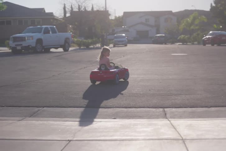 A happy young girl drives her red toy convertible car down the driveway and around her quiet suburban neighborhood street