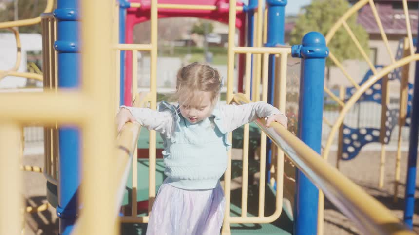 A little girl in a tutu smiles as she carefully walks across a colorful playground structure in slow motion