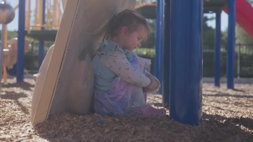 A young girl sits alone under a playground looking sad, then looks up and gives a sweet smile to the camera
