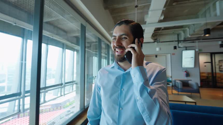 A smiling young businessman in a blue shirt enjoys a pleasant phone conversation while standing by a large window in a modern office