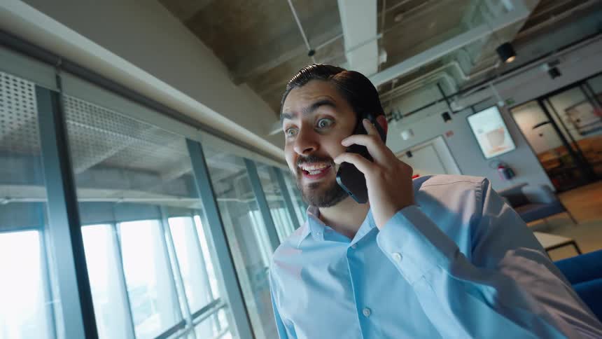 A young professional receives exciting news on a phone call while standing in a modern office lounge
