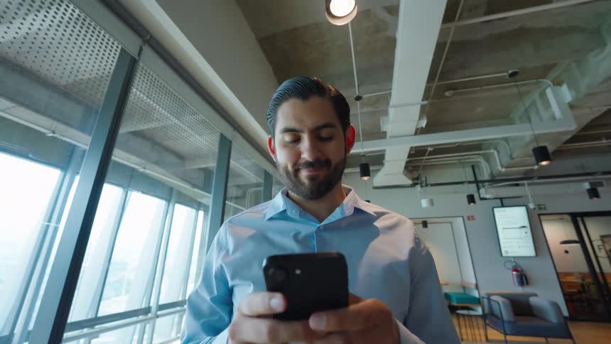 Low-angle dolly shot of a smiling businessman using his smartphone in a modern, open-plan office