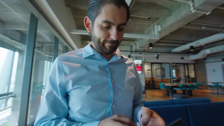 A handsome young businessman smiles while texting on his smartphone in a modern office lounge area