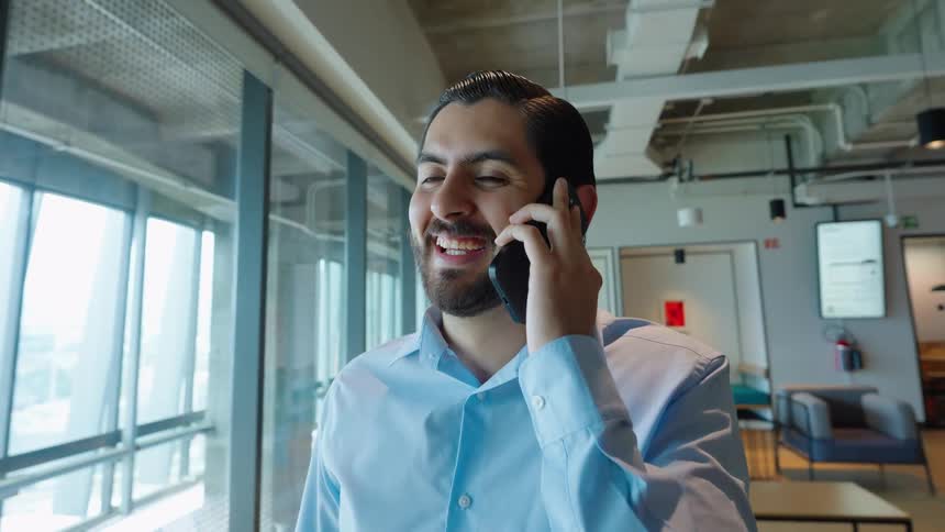 A happy businessman in a light blue shirt smiles while talking on his smartphone in a modern office space