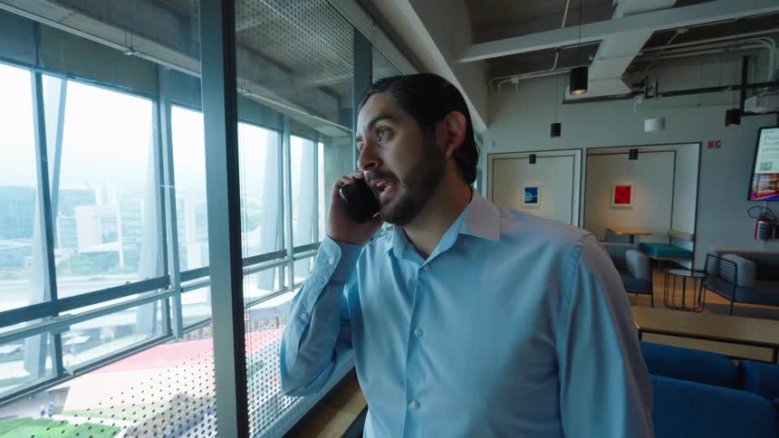 A smiling businessman's expression turns to concern while taking a serious phone call by a window in a modern office