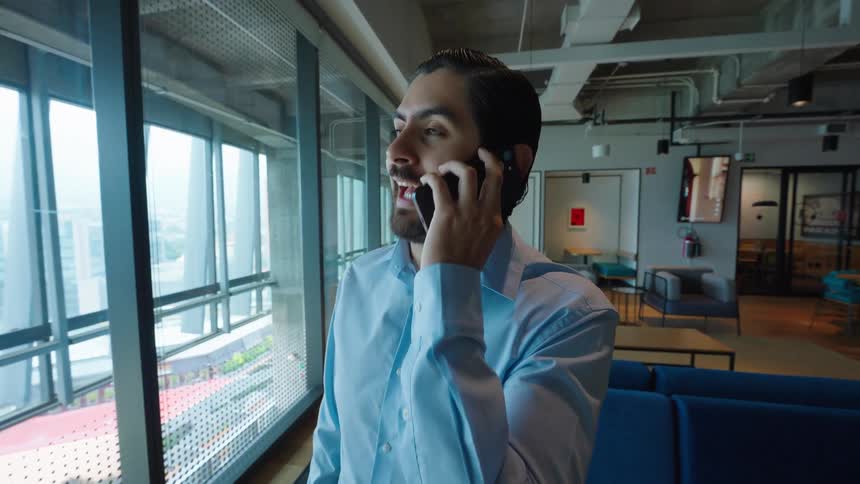 Happy young businessman talking on his smartphone while looking out a large window in a modern office