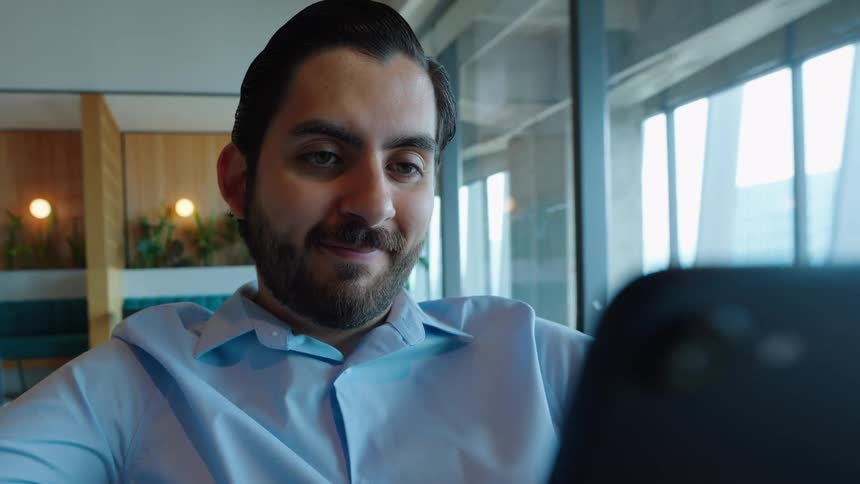 A smiling businessman in a blue shirt happily scrolls through his smartphone in a modern, brightly lit office lounge