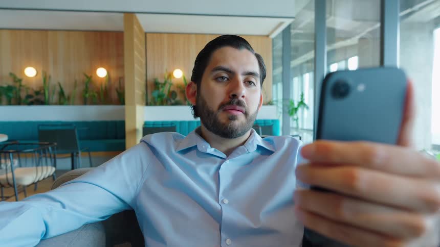 A businessman with a beard has a video call on his smartphone while relaxing in a modern office lounge