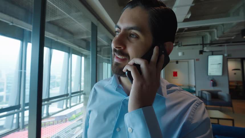 A young businessman in a modern office answers a phone call with a smile while looking out the window