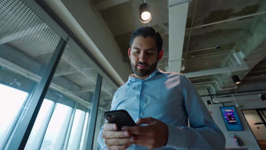 A handsome businessman finishes a call and then uses his smartphone in a modern, open-plan office space