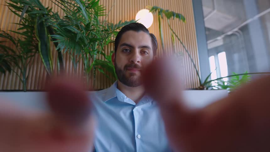 Point of view of a person using a mobile device, with a young businessman watching attentively in a modern, plant-filled setting