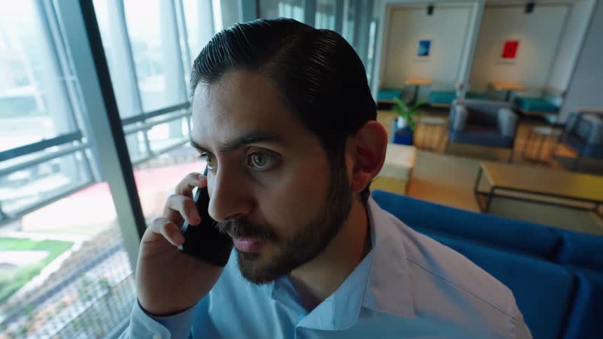 A handsome businessman in a modern office receives shocking news during a phone call by the window