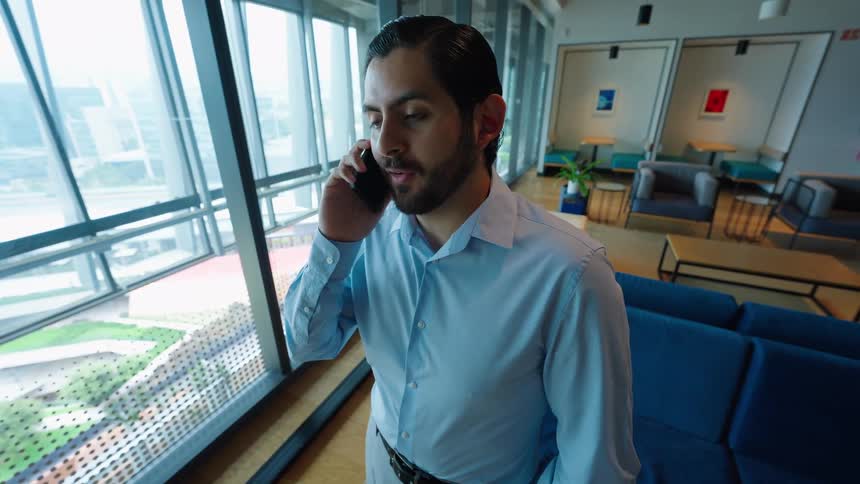 A well-dressed businessman engages in a phone conversation while standing by a large window in a modern office lounge
