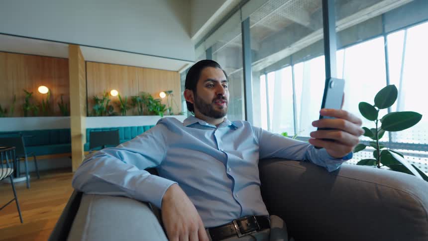 A well-dressed businessman engages in a video call on his smartphone while relaxing in a modern office lounge