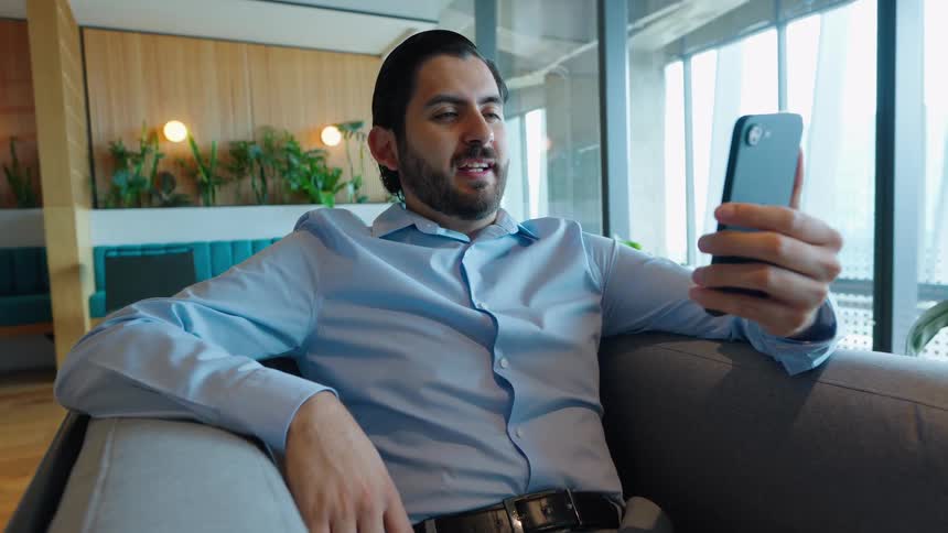 A businessman sits comfortably in a modern office lounge while having an animated video call on his smartphone