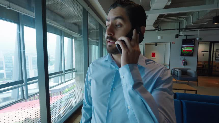 A young businessman in a modern office answers his phone and receives surprising good news while looking out the window