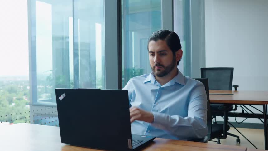 Pensive businessman in a modern, glass-walled office stops typing on his laptop to contemplate and look outside