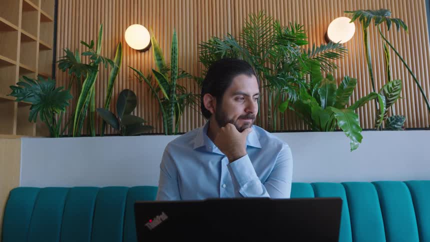 A thoughtful bearded man works on his laptop, deep in concentration in a modern, stylish office space with green plants