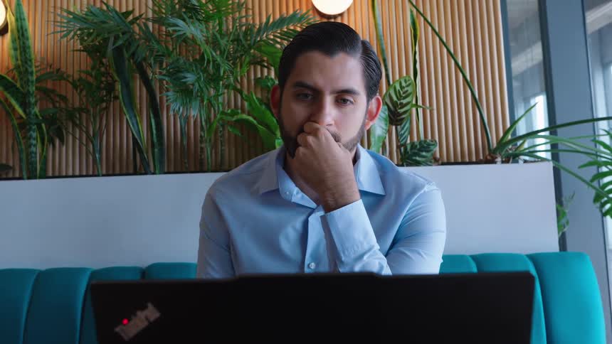 A pensive businessman in a blue shirt looks thoughtfully at his laptop while working in a modern office lounge