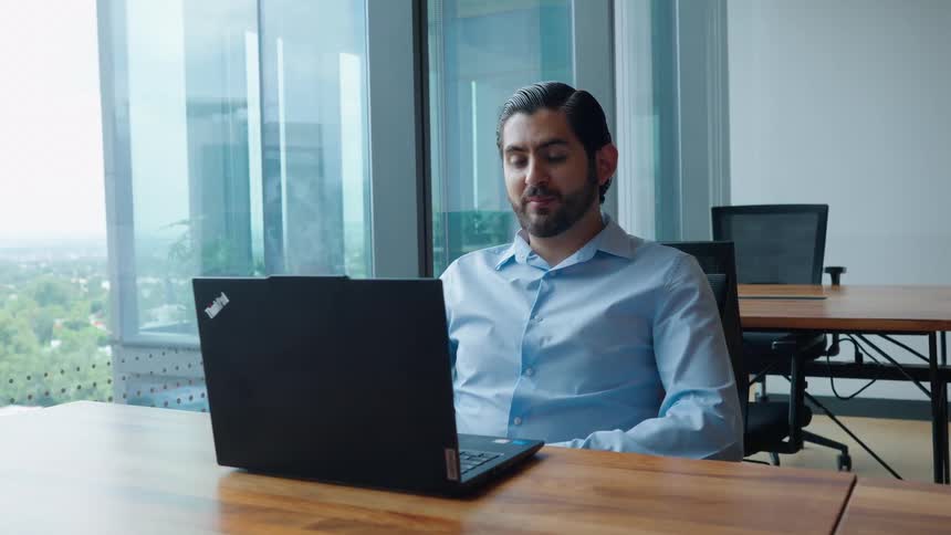 A professional businessman sits at a desk in a modern office and engages in a video call on his laptop