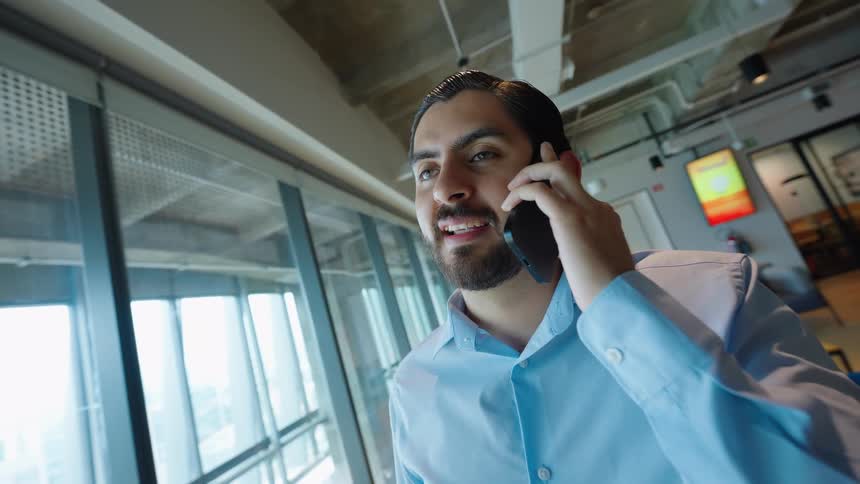 A happy young businessman smiles while talking on his smartphone in a modern, sunlit office