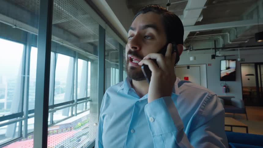 A businessman with a beard talks on his smartphone while standing by the window in a modern office
