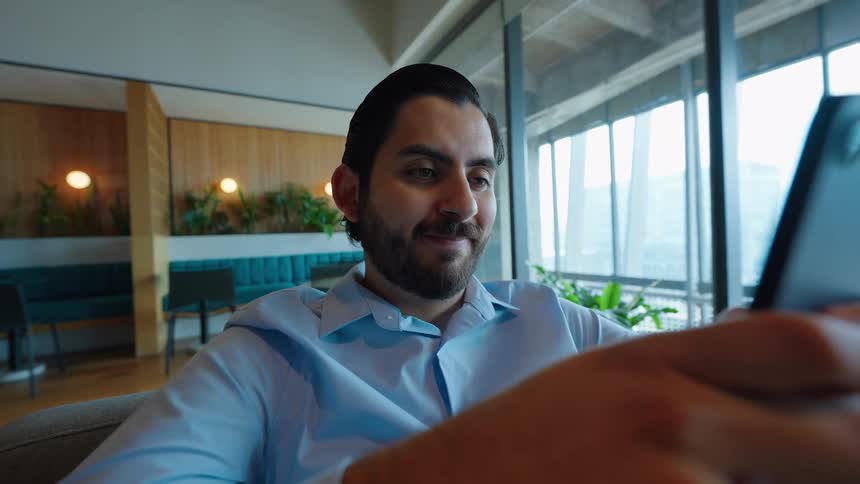 A smiling businessman sits in a modern office lounge and texts on his smartphone during a break