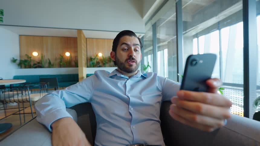 A man in a modern office lounge reacts with shock and surprise while on a video call on his smartphone