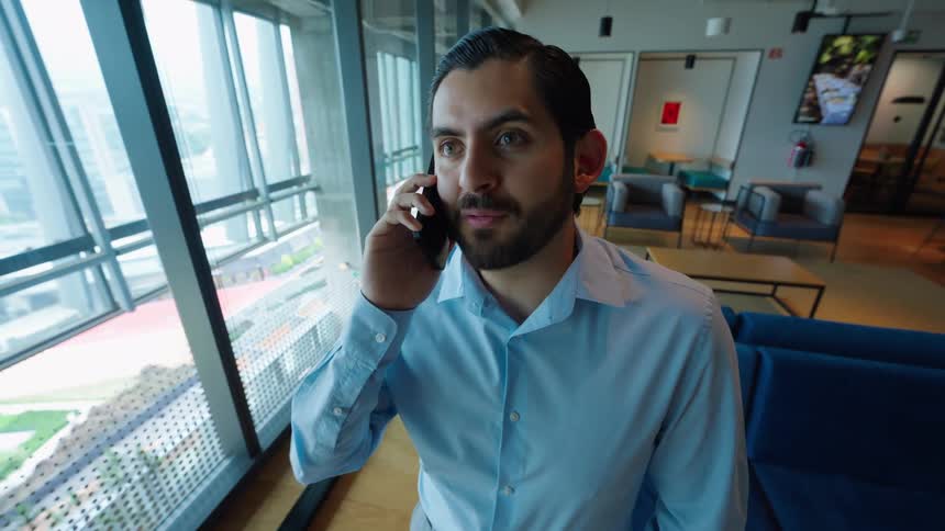 A businessman reacts with shock and disbelief while on a phone call, standing by the window in a modern office