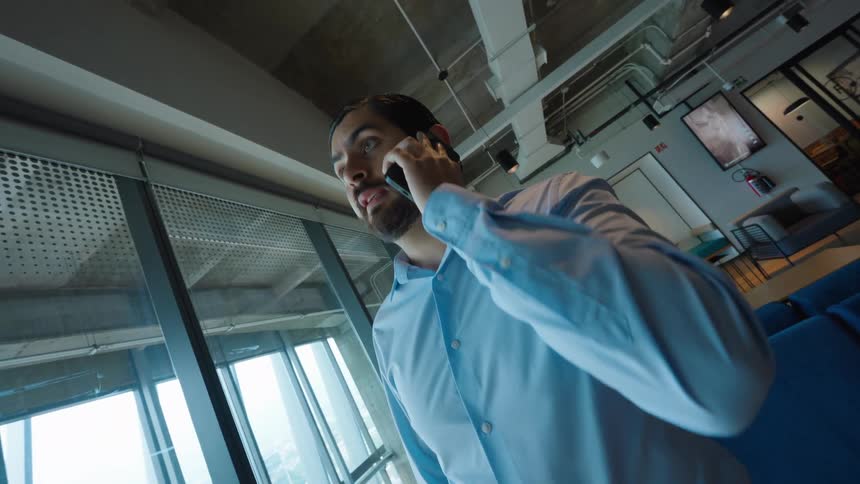 A serious businessman stands near a large window in a modern high-rise office while talking on his smartphone