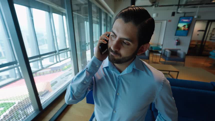 A young businessman in a modern office answers a phone call while standing next to a large window