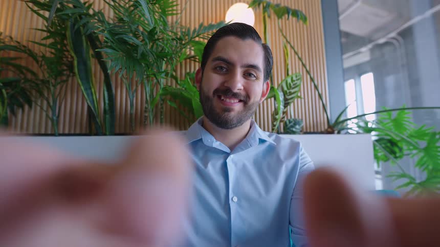 POV shot of a kind, bearded man smiling and reaching out for a warm hug in a modern, plant-filled office