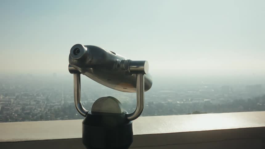 A coin-operated binocular viewer on an observatory deck overlooks a vast, hazy cityscape
