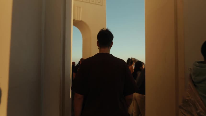 Following a young man in slow motion as he walks through the sunlit colonnade of a landmark building crowded with tourists