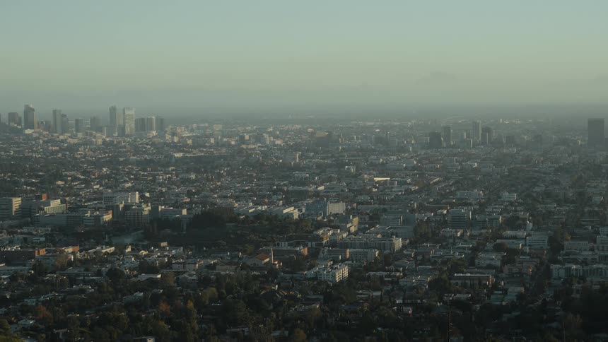 A vast and sprawling cityscape of Los Angeles is seen from a high angle during a hazy golden hour, with downtown skyscrapers in the distance