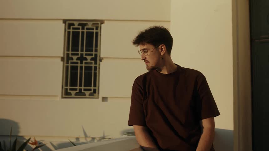 A pensive young man in glasses watches the day turn to night in a time-lapse from his balcony