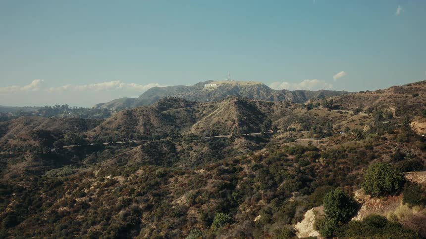 A scenic wide shot of the iconic Hollywood sign perched atop the arid, brush-covered Hollywood Hills on a clear, sunny day in Los Angeles