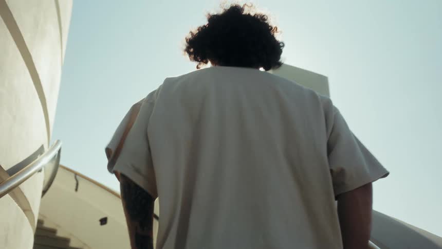 Low angle view of a curly-haired man walking up a bright, sunlit staircase in slow motion