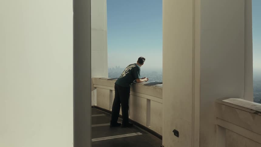 A young man walks onto a sunlit balcony, framed by white columns, to gaze out at the vast and hazy city skyline below