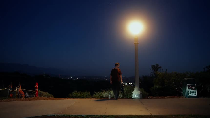 A young man playfully dances and leans against a bright lamppost at night on a scenic overlook with a city skyline in the distance