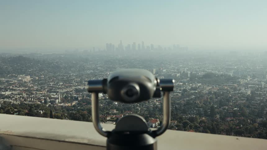 An observation binocular viewer in the foreground overlooks the vast, hazy cityscape of Los Angeles in the distance